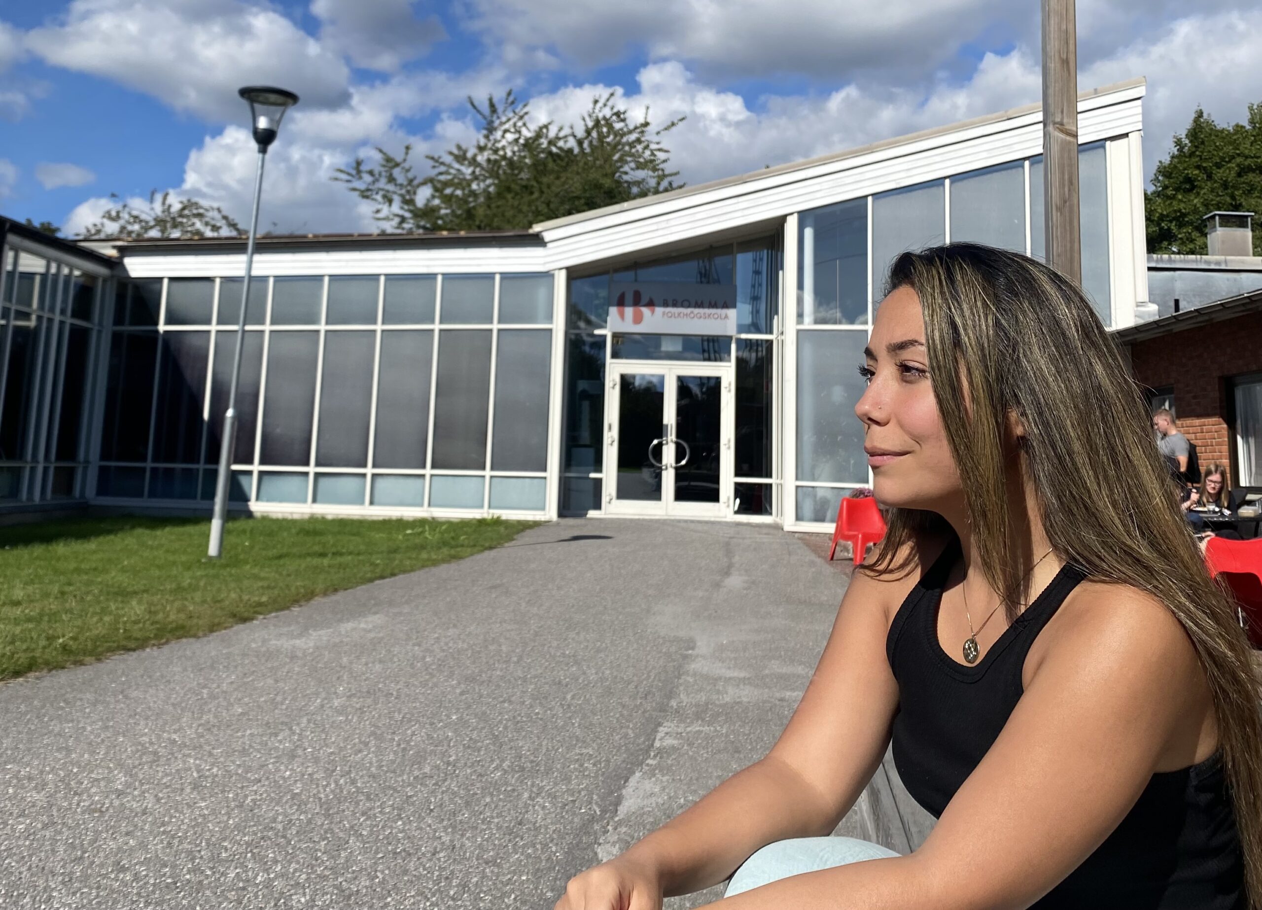 Kvinnlig studerande sitter i solen utanför skolans huvudentré